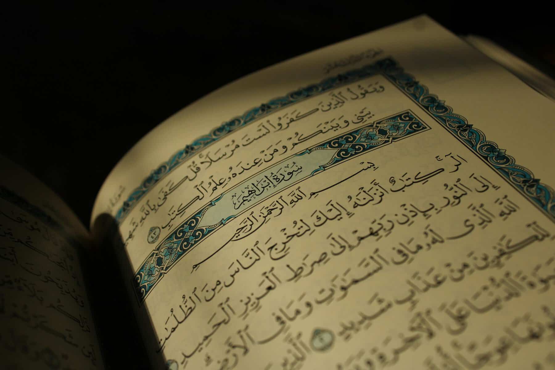 Open Mushaf showing the start of Surah Ibrahim with ornate blue cartouche, lit against a black background