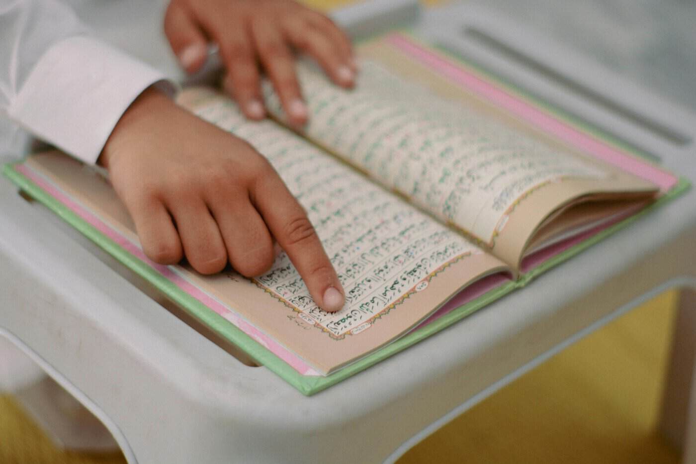 Close-up of a child's hands on an open Mushaf on a small white desk, with a finger tracing along the Arabic text, the classical hifz memorisation technique