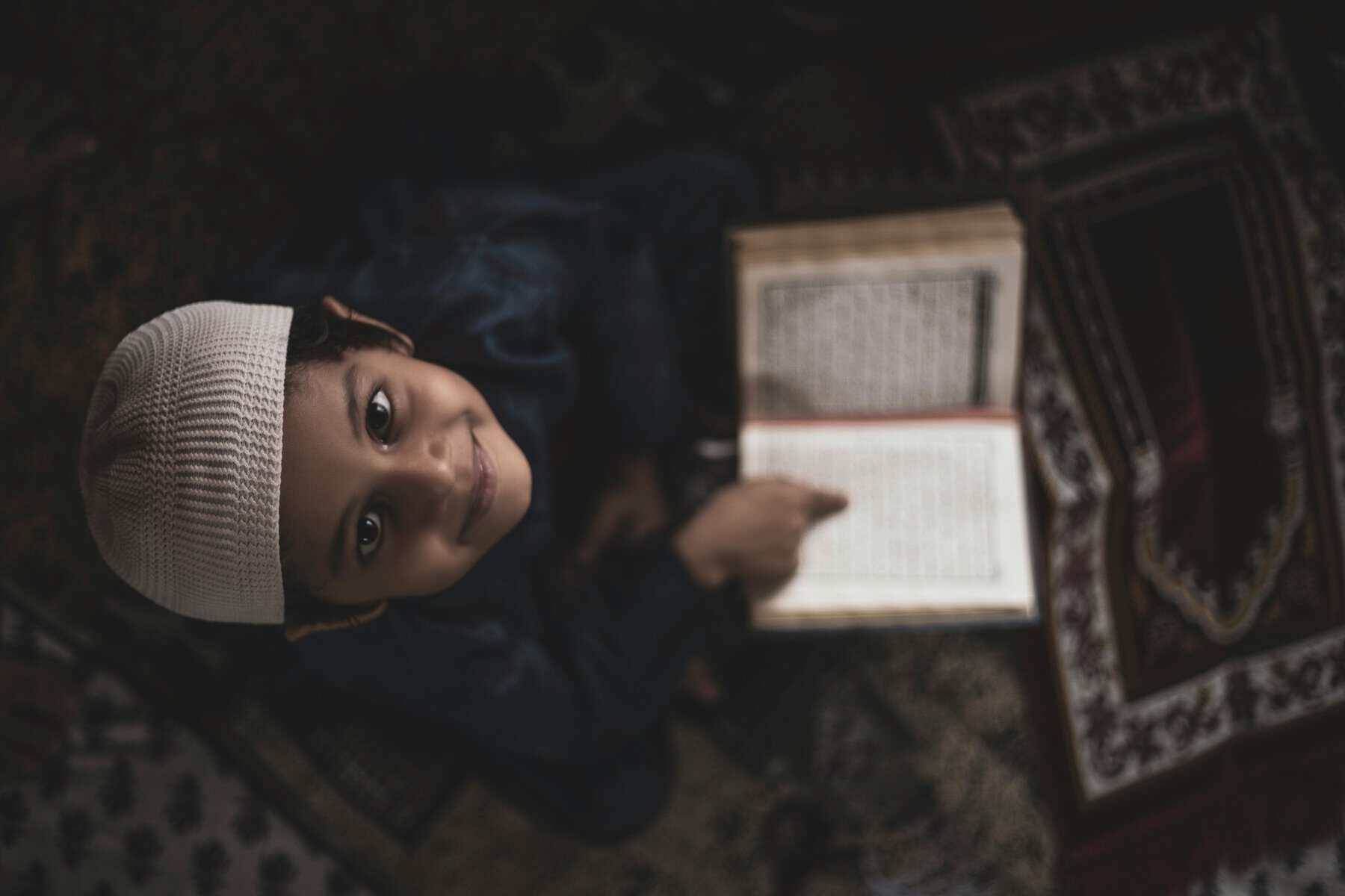 A young Muslim boy in a white skull cap and dark thawb kneeling on a patterned prayer mat with an open Mushaf in front of him, looking up and smiling