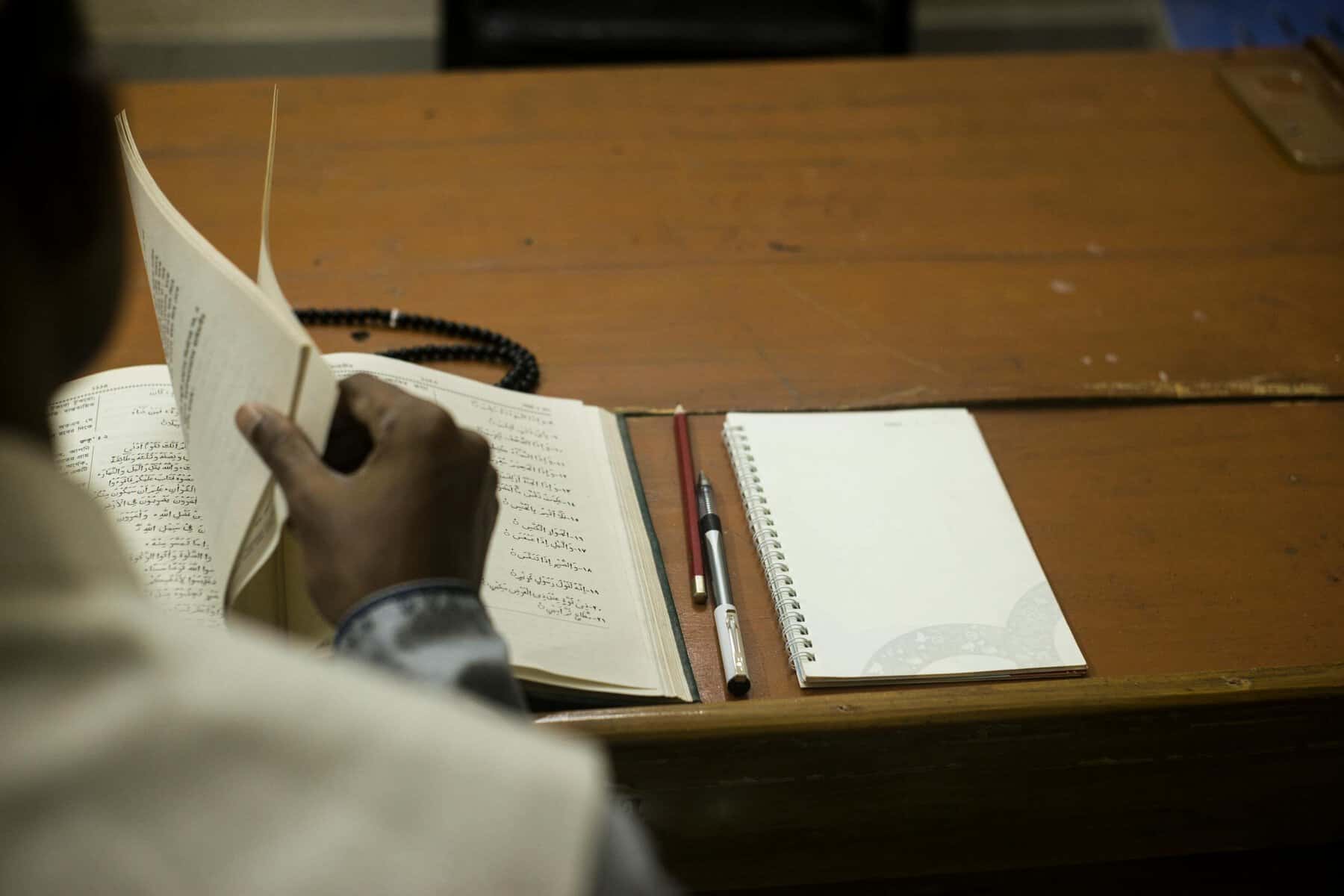 Real student in white thawb studying an open Islamic fiqh text with Arabic rulings on a wooden desk, with prayer beads and a notebook ready for study