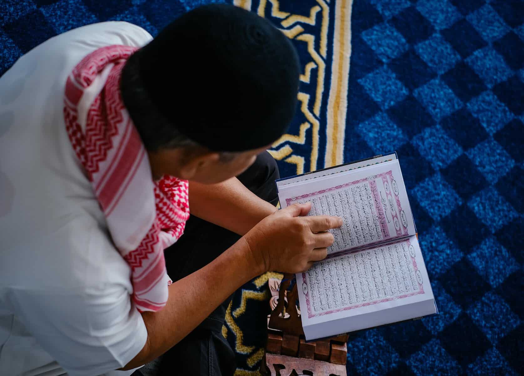 A Muslim man in a keffiyeh and skull cap pointing at the Arabic text of an open Mushaf placed on a wooden rehal stand, kneeling on a blue prayer rug