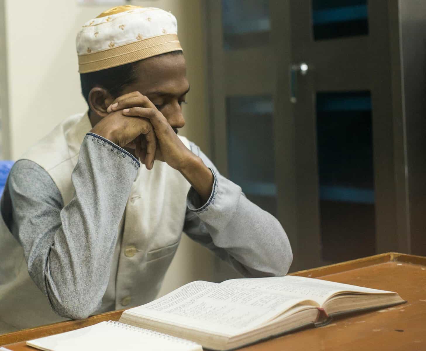Young Muslim man in white skullcap and waistcoat memorising Quran at a wooden desk with notebook beside him