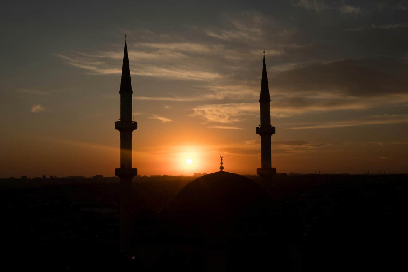 A mosque with two minarets and a central dome silhouetted against an orange sunset sky, evoking the quiet anticipation of the moment Ramadan begins