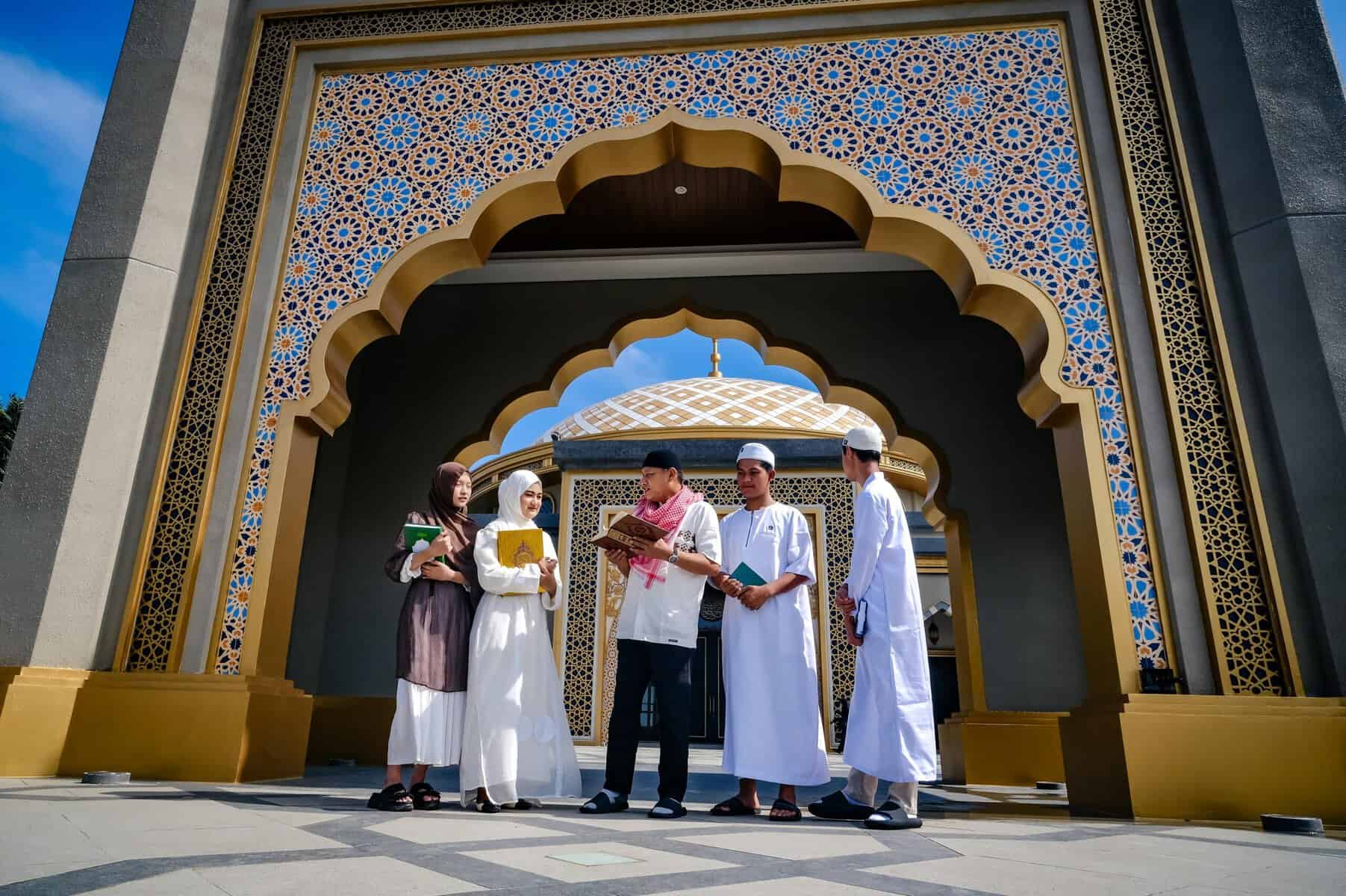Five young Muslim students reading and discussing Quran texts at the geometric-tiled arched entrance of a domed mosque