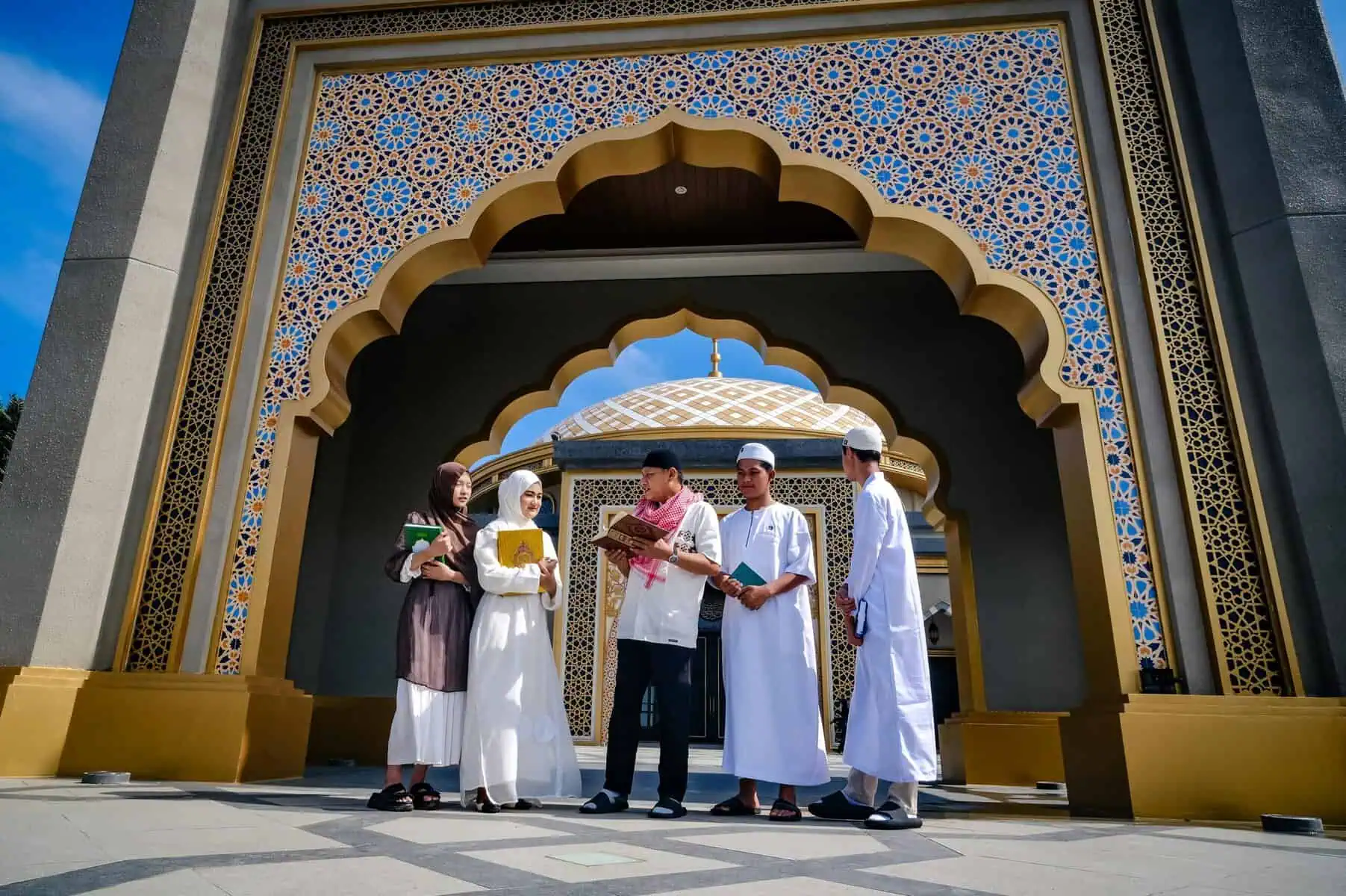 Five young Muslim students reading and discussing Quran texts at the geometric-tiled arched entrance of a domed mosque