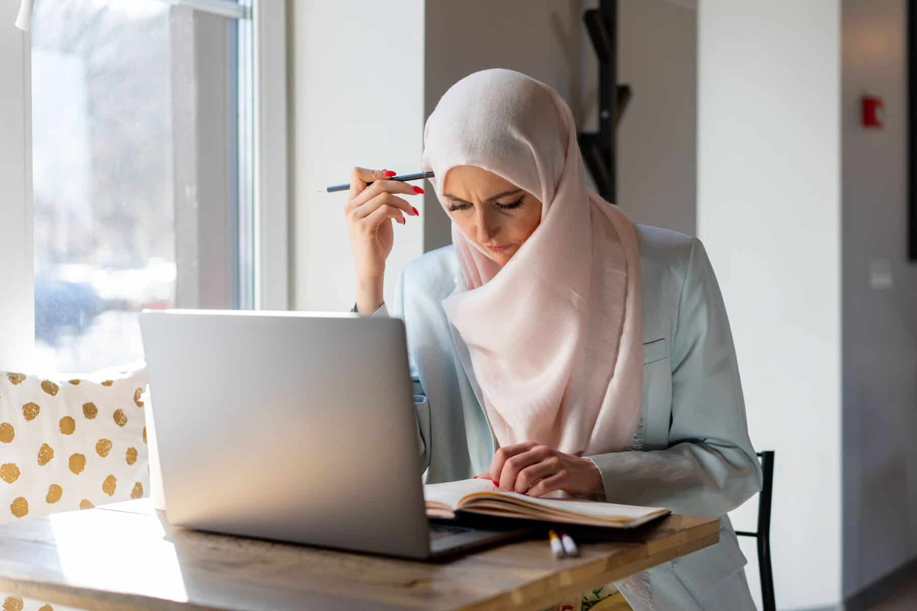 Muslim woman in pale pink hijab focused on her laptop and notebook at a sunlit home desk