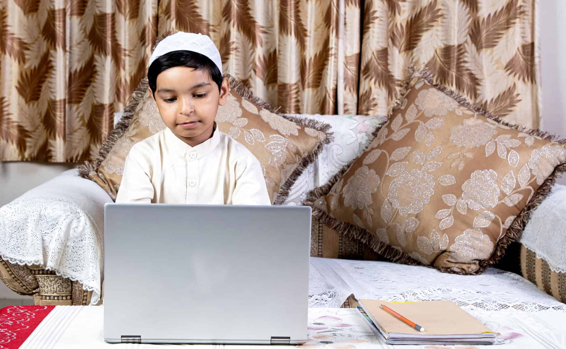 A young Muslim boy in a white thawb and skull cap sitting on a couch, focused on his laptop during an online Quran class at home in the UK