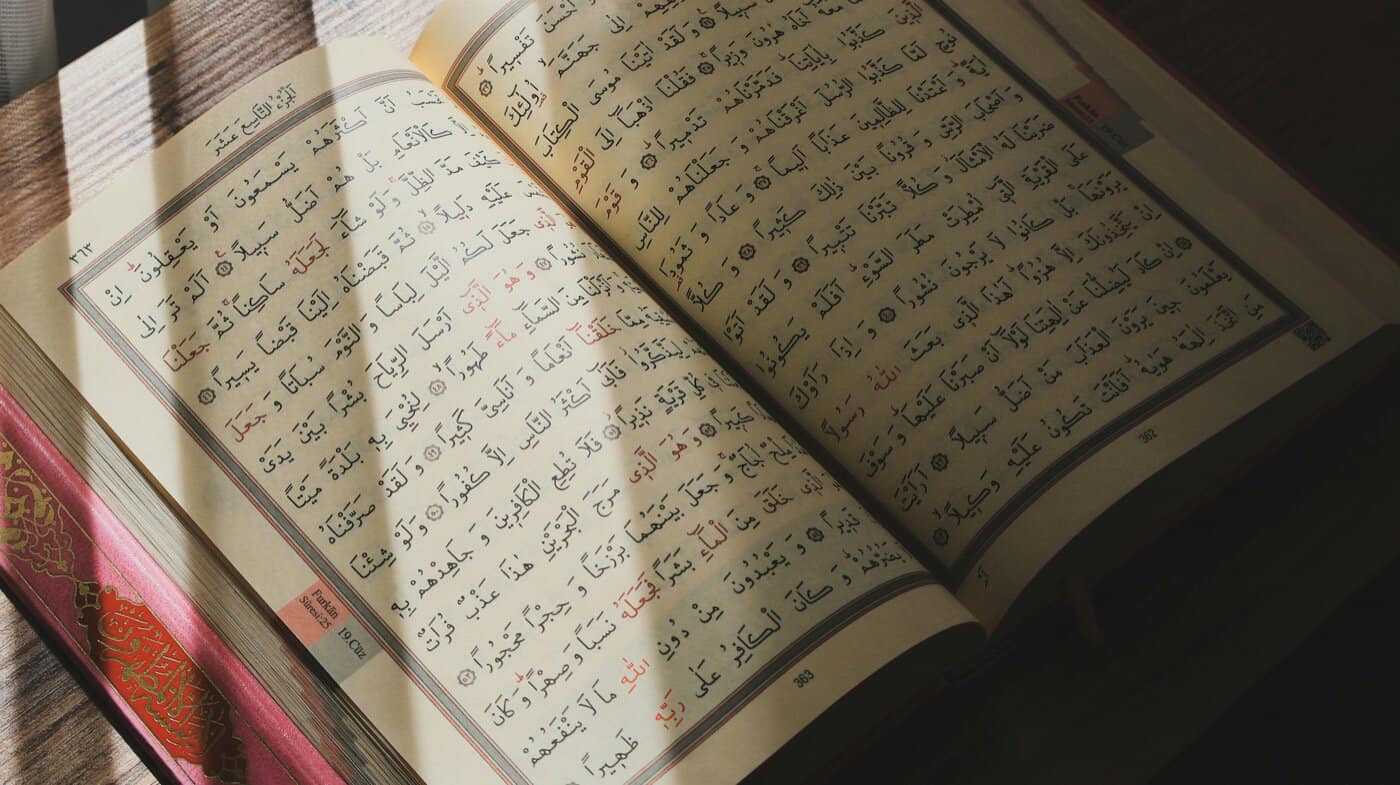Close-up of an open Mushaf showing the Arabic text of the Qur'an with red diacritical marks and a decorative gold and pink border, lit by warm sunlight on a wooden surface