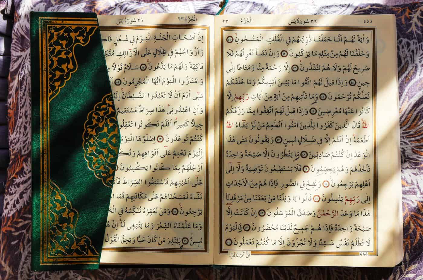 An open Mushaf displaying the Arabic text of Surah Ya-Sin with an ornate green and gold cover, lit by warm sunlight