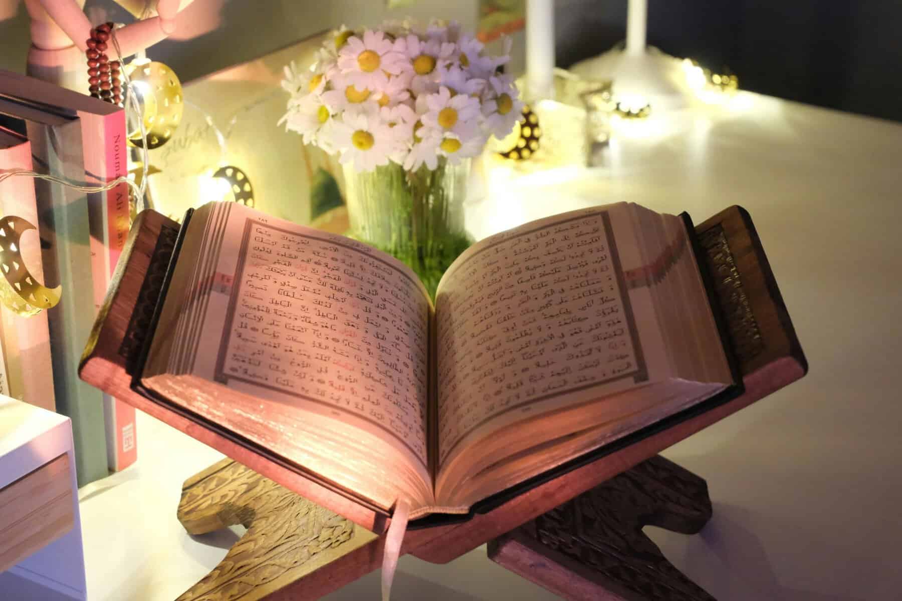Open Mushaf on a carved wooden rehal stand at home, lit by warm string fairy lights with a vase of white daisies behind