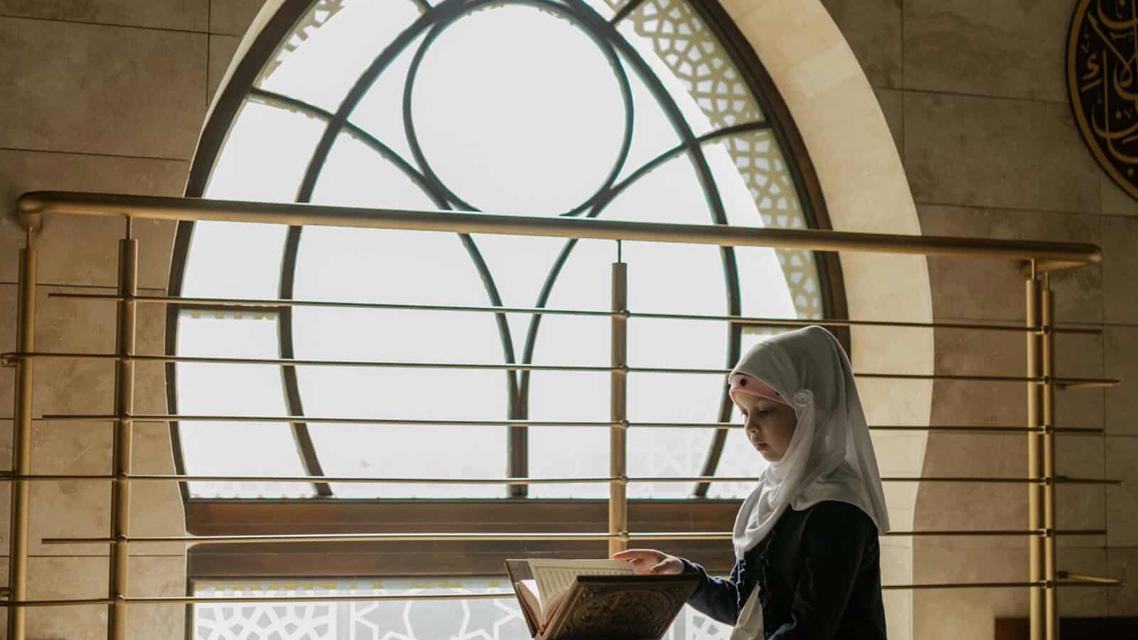 Woman reading the Quran on a rahl in front of a large arched mosque window