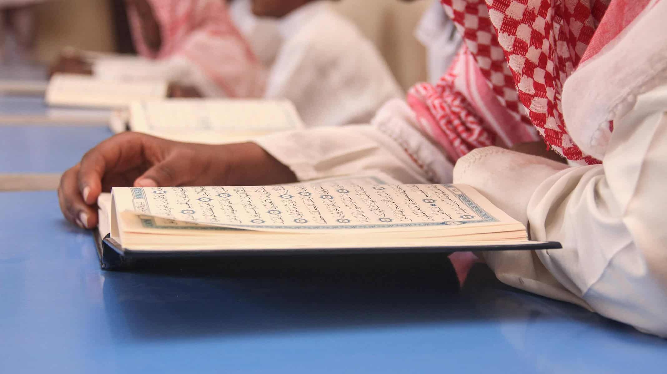 Students in traditional attire reading from open Mushafs at a study desk, illustrating seeking knowledge in Islam through classical Quran study