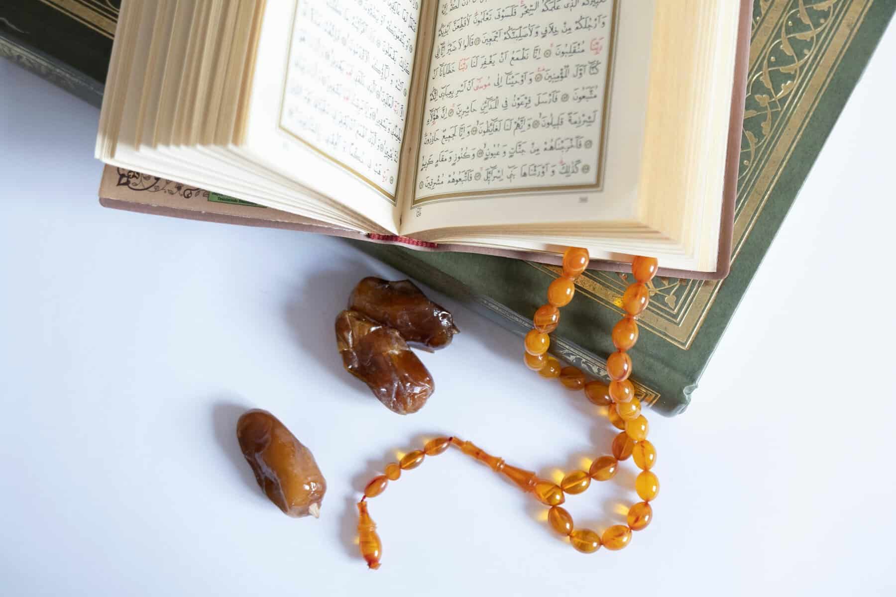 An open Mushaf displaying Arabic verses, an amber tasbih, and three dried dates on a clean white surface, the three core preparation tools for the month of Ramadan