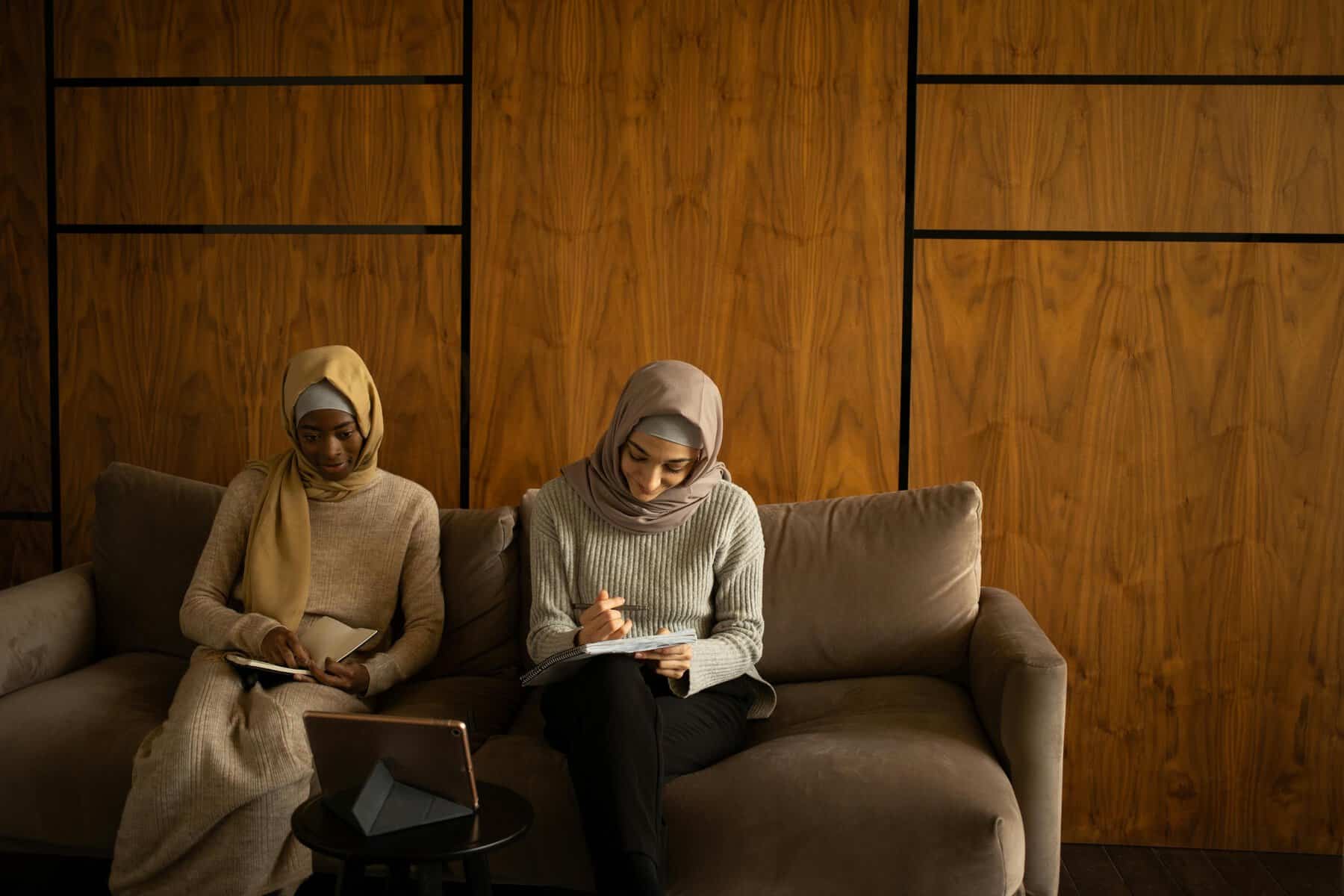 Two Muslim students in hijabs sitting on a couch with notebooks and a laptop, engaged in a structured Islamic studies session at home