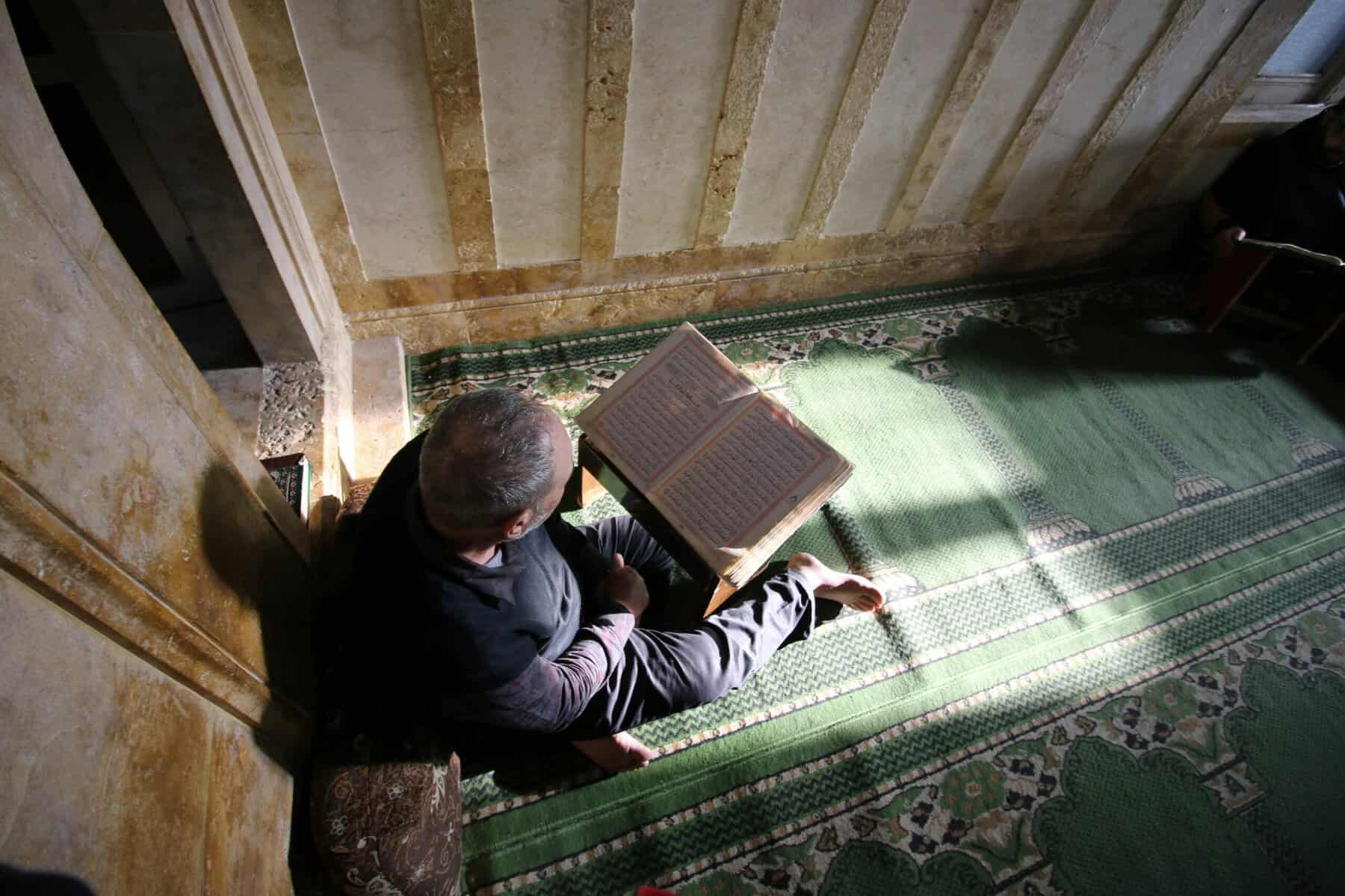 Adult man in dark robes seated on a patterned green prayer carpet reading an open Mushaf on a wooden rehal inside a traditional Syrian mosque with striped ablaq stonework