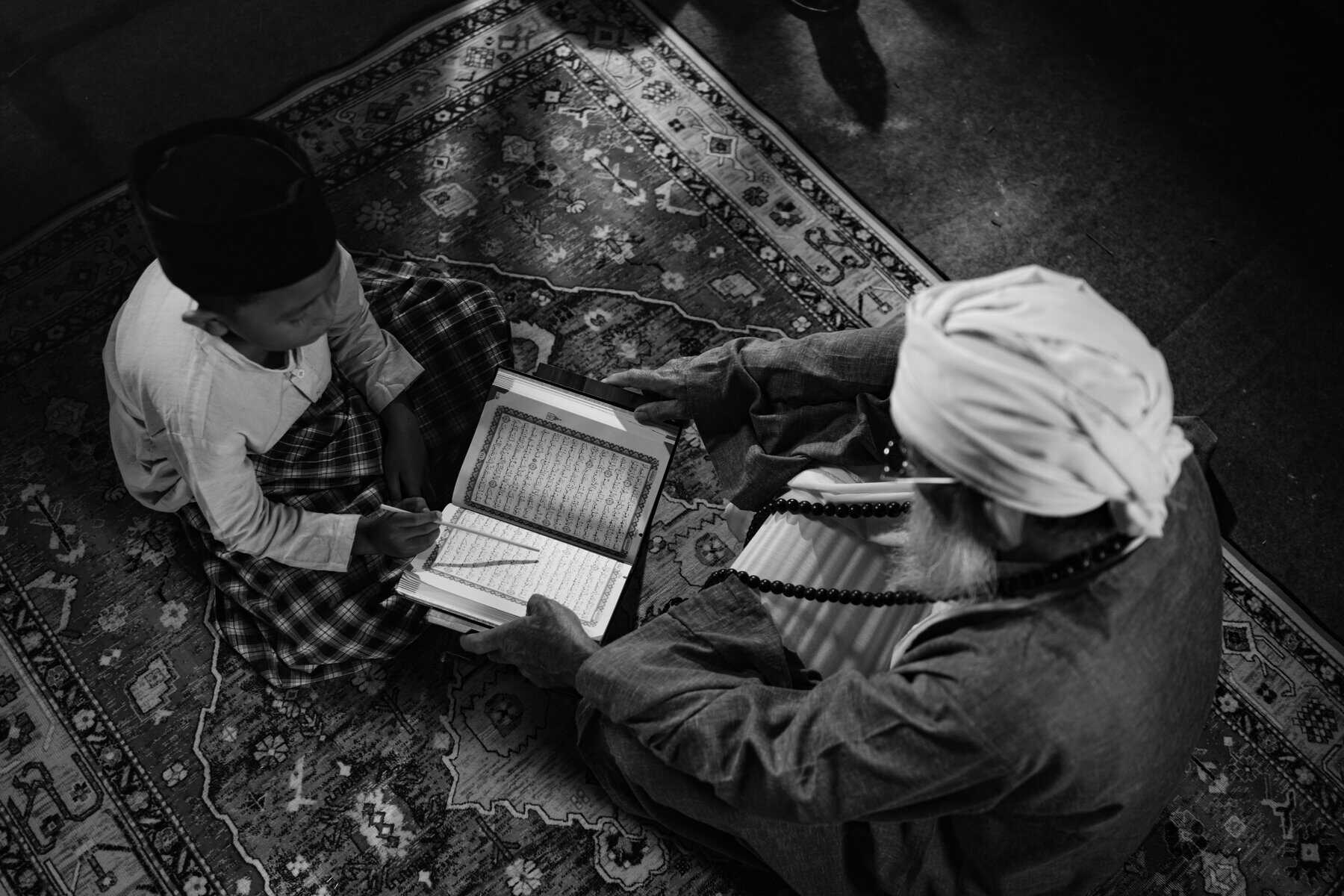 Black and white overhead shot of a young boy in white skullcap concentrating on an open Mushaf with an elder in white turban holding tasbih beside him on an ornate carpet