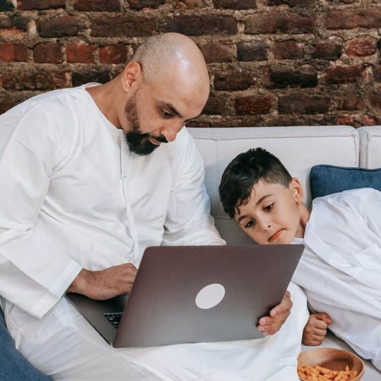 A father and son in traditional clothing taking an online class together on a laptop at home