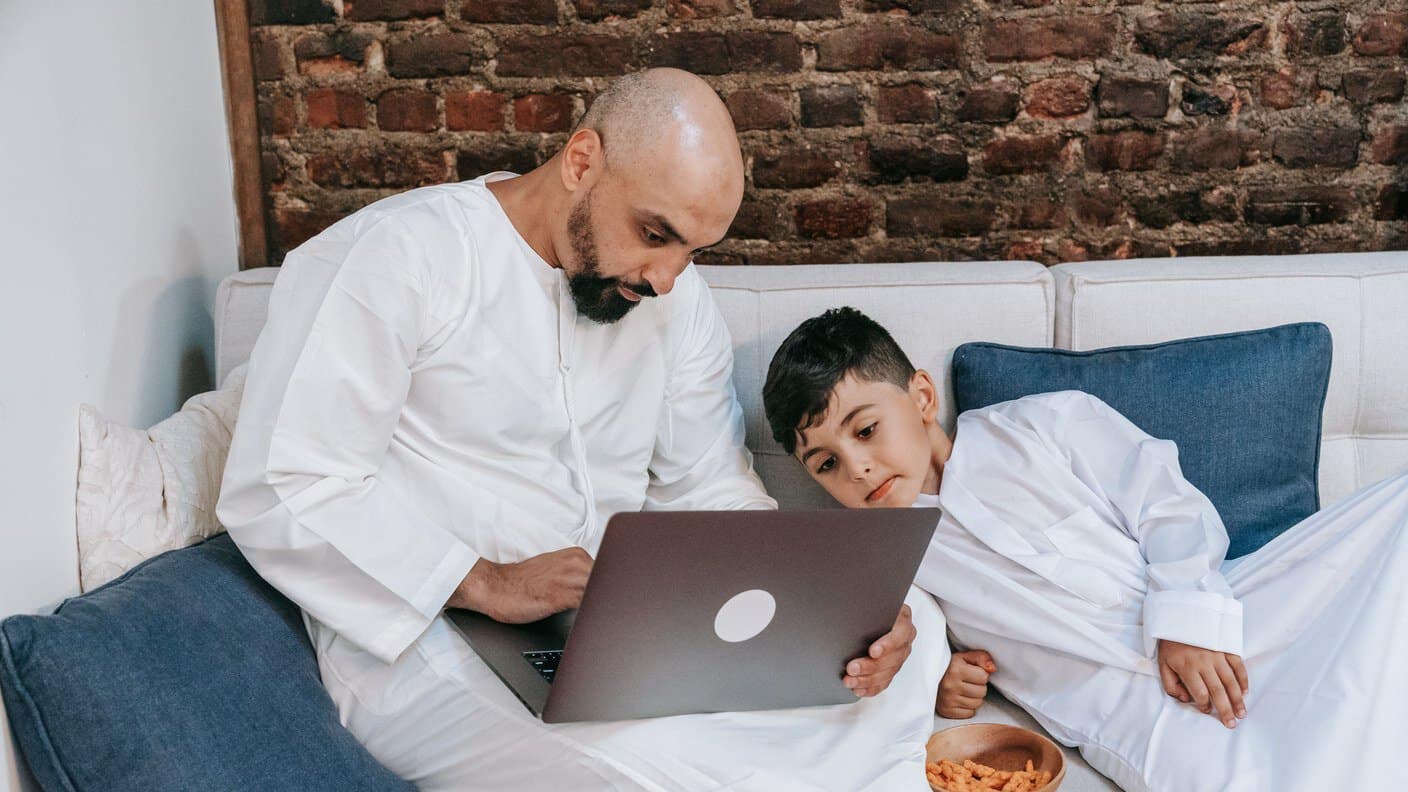 A father and son in traditional clothing taking an online class together on a laptop at home