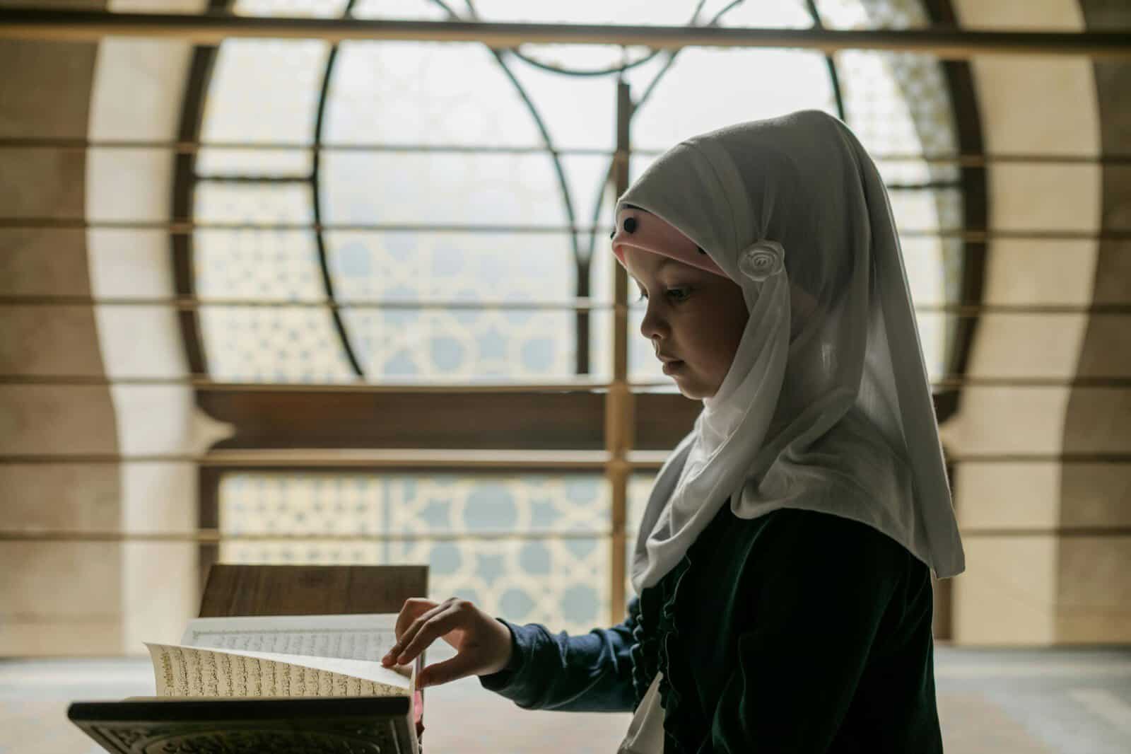 A young girl in hijab reading the Quran from a wooden stand, a beginner learning to read