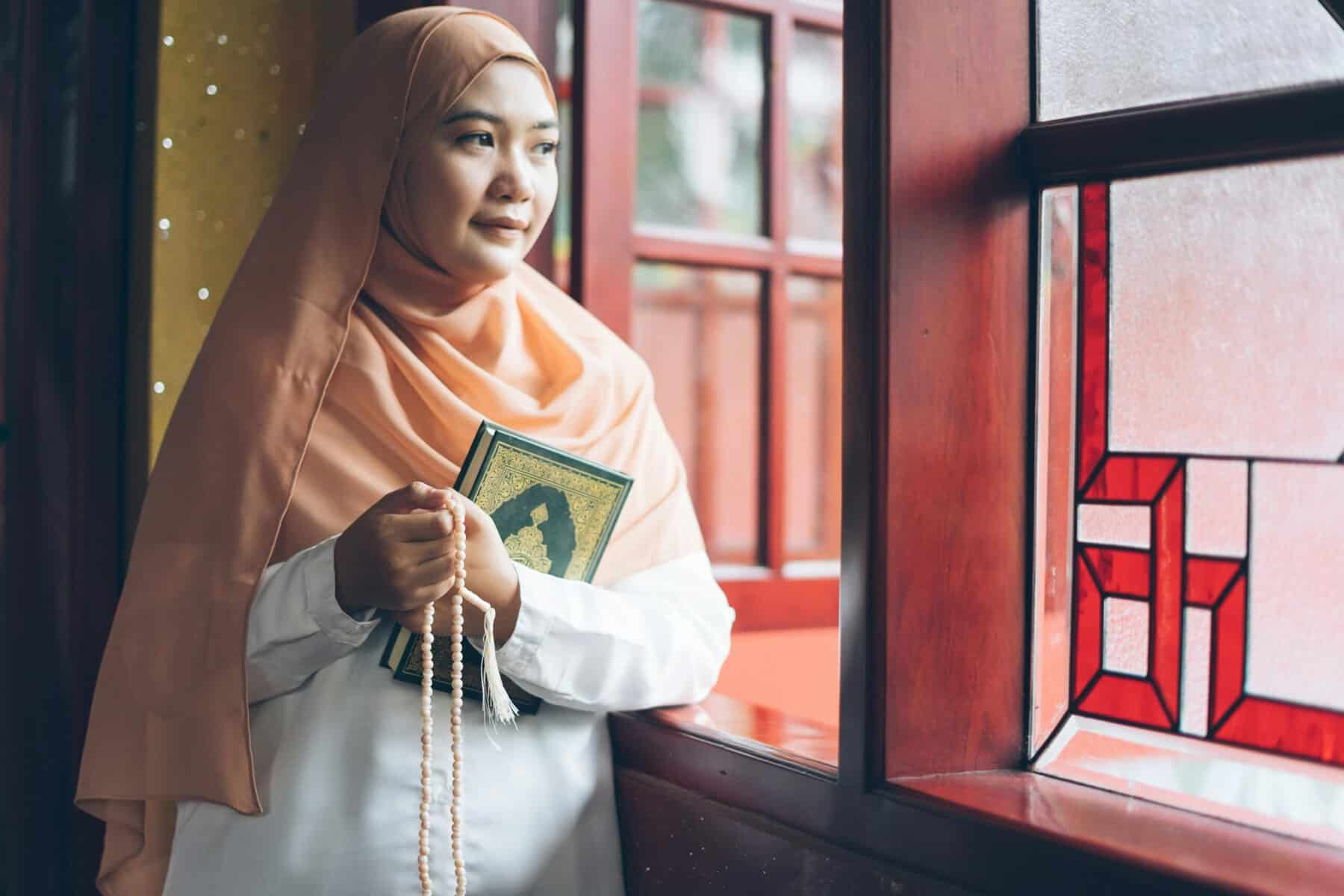 Adult Muslim woman in a peach hijab holding a green and gold-embossed Mushaf and tasbih prayer beads, standing by a red-framed stained-glass window