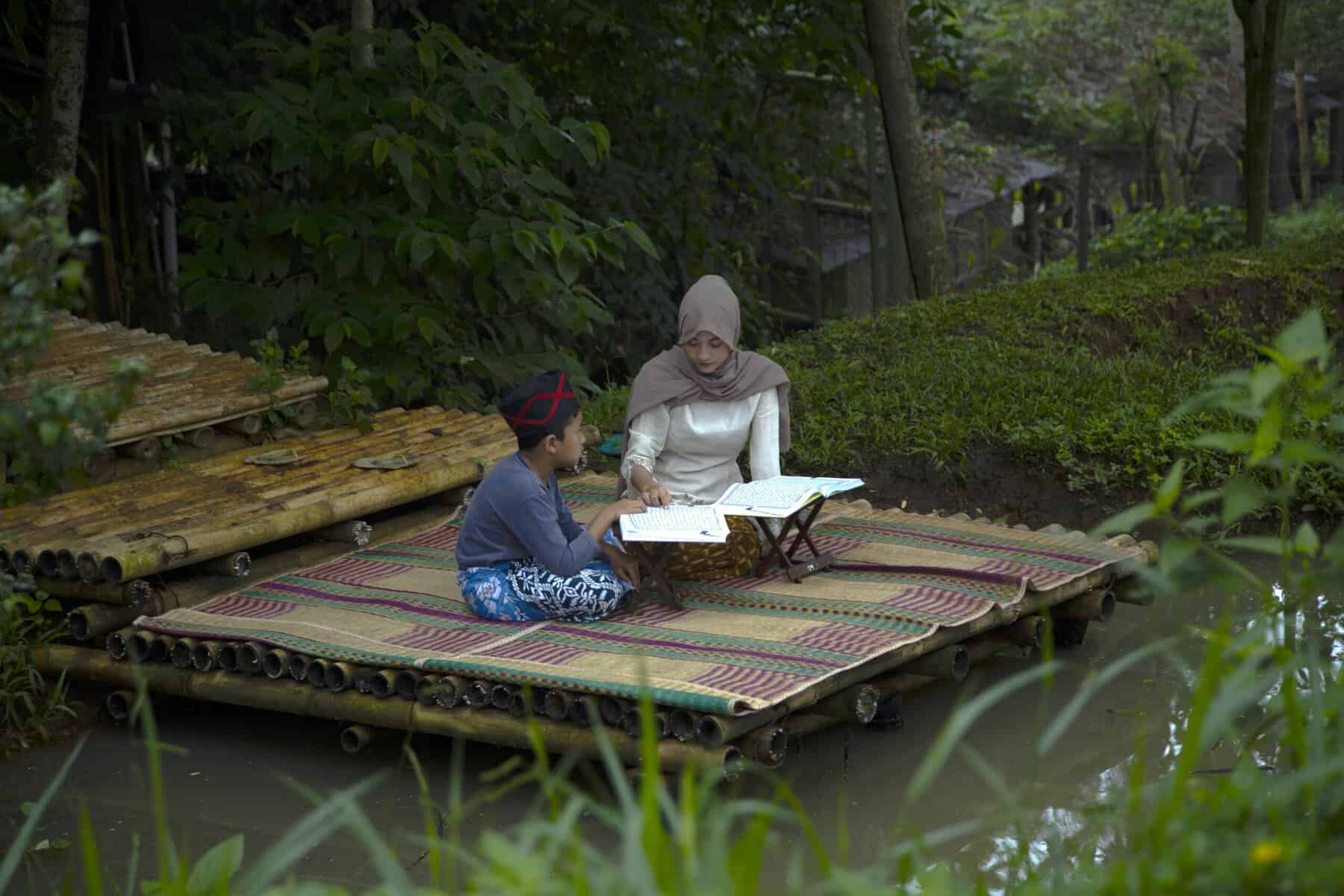 Young Muslim woman in tan hijab and a boy in skullcap seated cross-legged on a woven bamboo platform with an open Mushaf on a wooden rehal between them, surrounded by lush tropical greenery