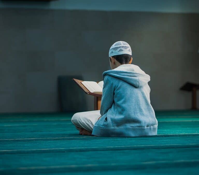 A young boy sitting on a mosque carpet reading the Quran from a wooden stand, beginning his Hifz journey