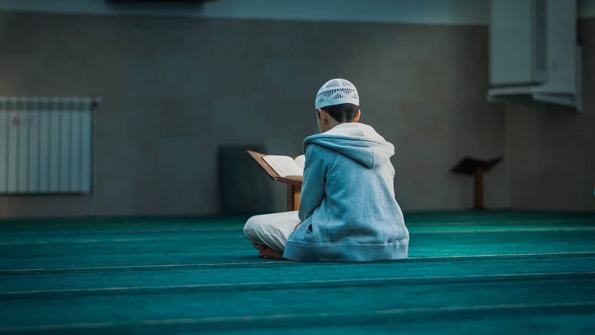 A young boy sitting on a mosque carpet reading the Quran from a wooden stand, beginning his Hifz journey