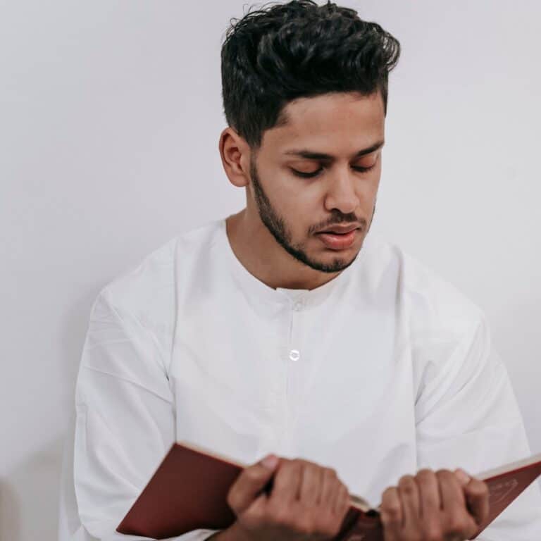A young man in a white thobe reciting from a Mushaf alone at home