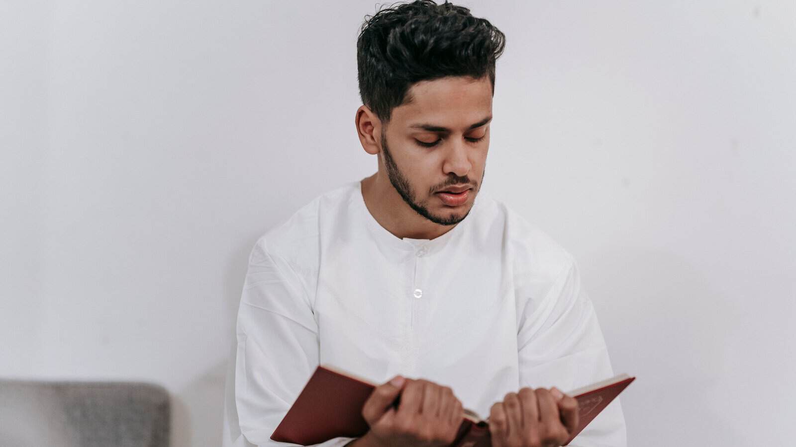 A young man in a white thobe reciting from a Mushaf alone at home