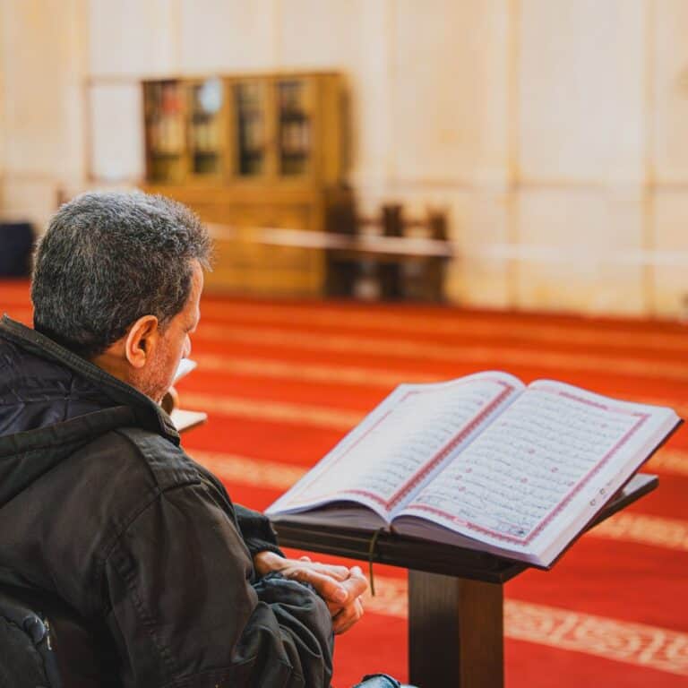 A man reciting from an open Mushaf on a stand in a mosque, pausing over the words