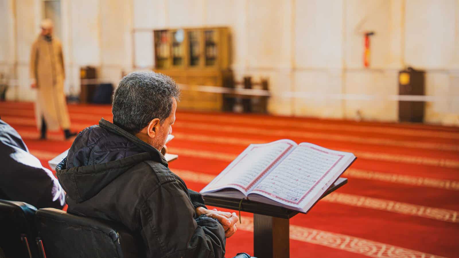 A man reciting from an open Mushaf on a stand in a mosque, pausing over the words