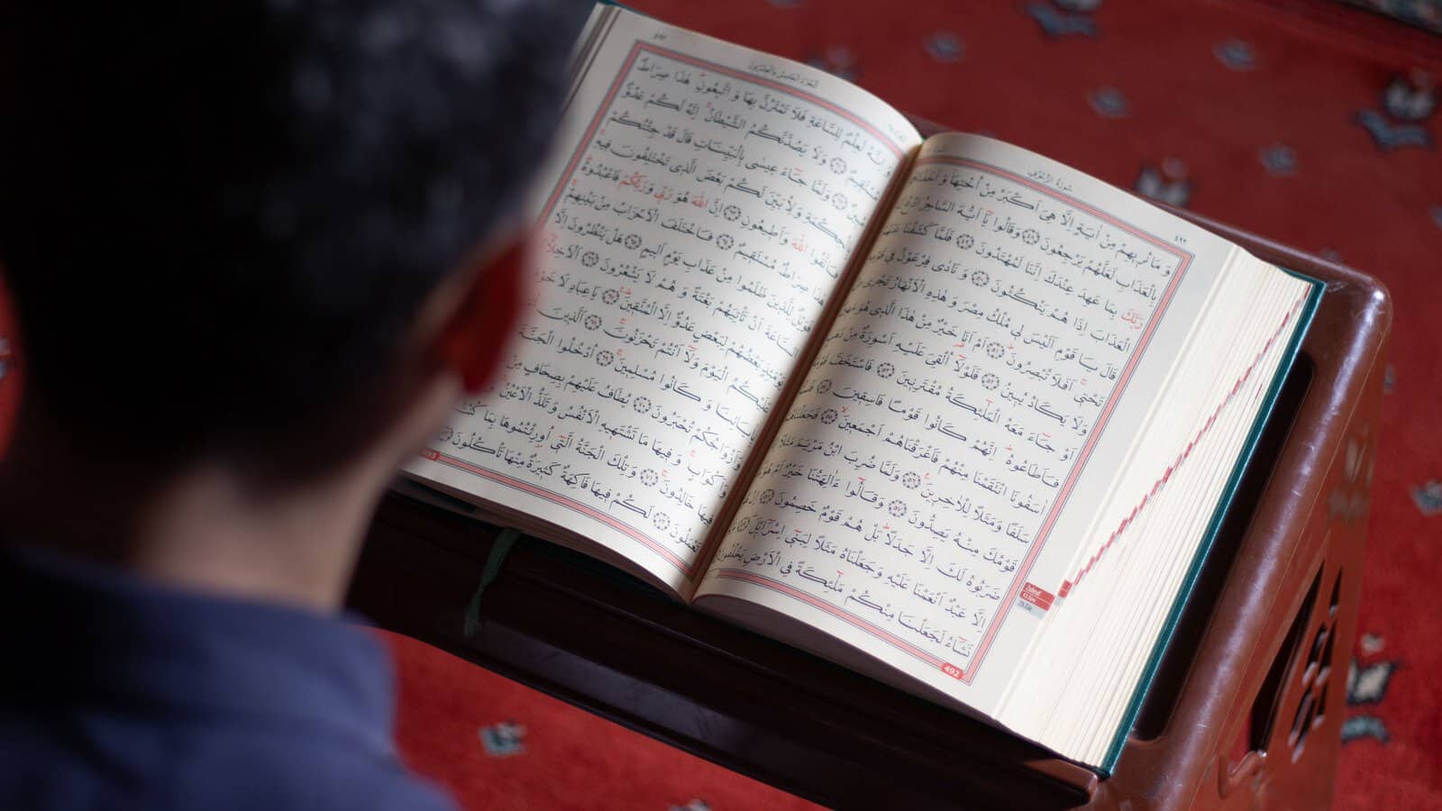 A man reading an open Mushaf on a wooden rehl stand in a mosque, viewed over his shoulder