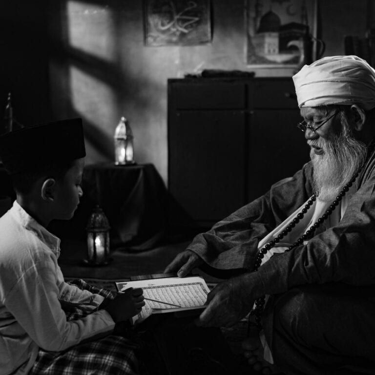 An elderly Quran teacher and a young boy reciting together from an open Mushaf, illustrating the one-to-one transmission behind an ijazah