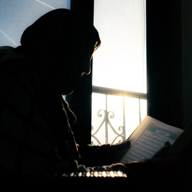 Silhouette of a woman in hijab reading the Quran by a window at sunset