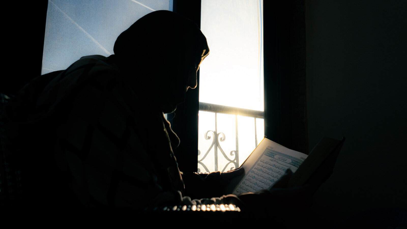 Silhouette of a woman in hijab reading the Quran by a window at sunset