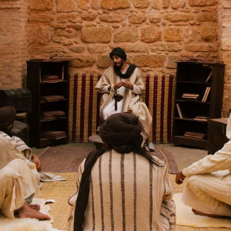 A teacher seated before shelves of manuscripts explaining a text to four seated students