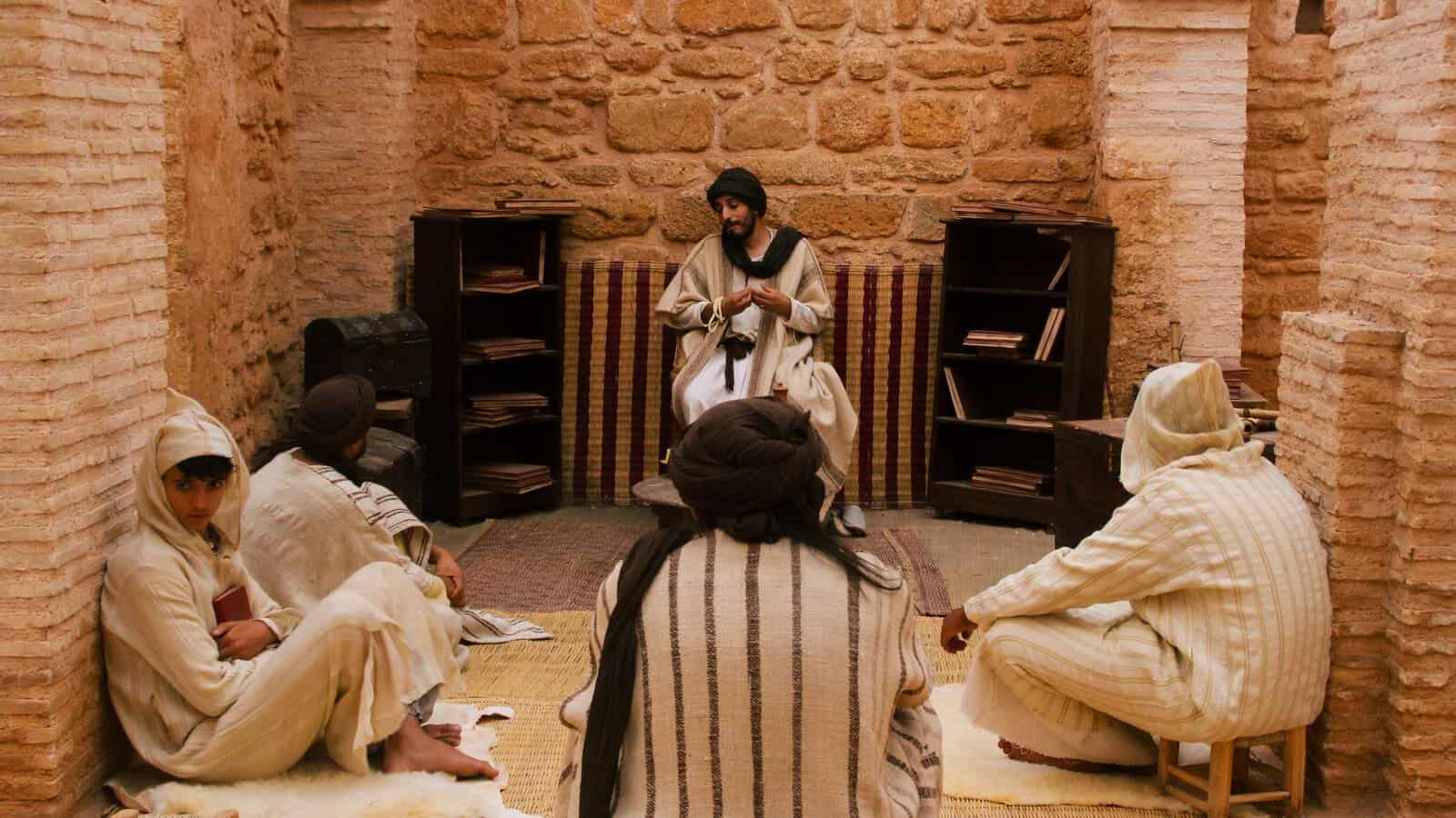 A teacher seated before shelves of manuscripts explaining a text to four seated students