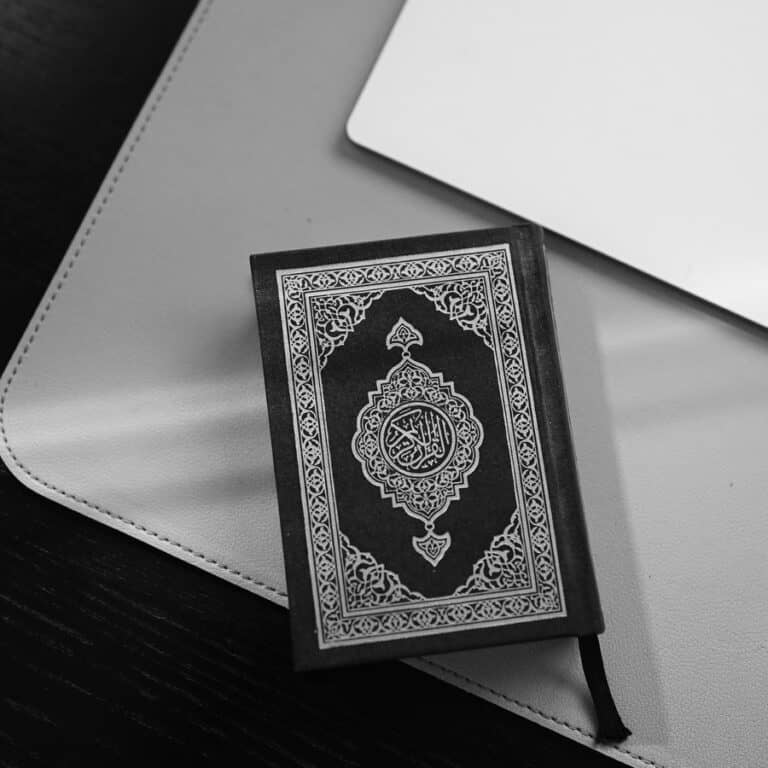 A small Quran with an ornate cover resting on a desk pad beside a keyboard and trackpad