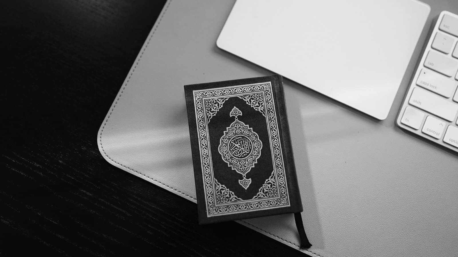 A small Quran with an ornate cover resting on a desk pad beside a keyboard and trackpad