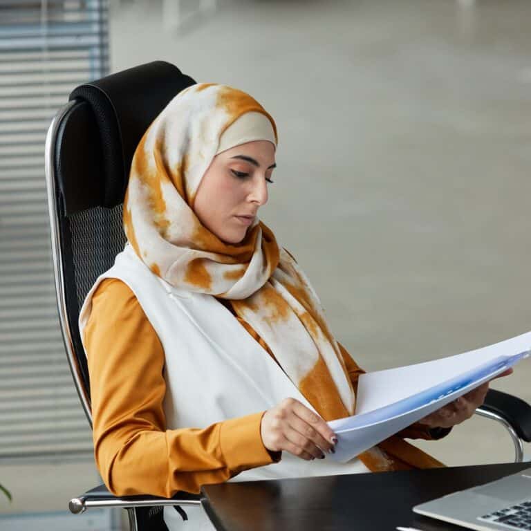 A woman in a hijab carefully reviewing documents at a desk with a laptop