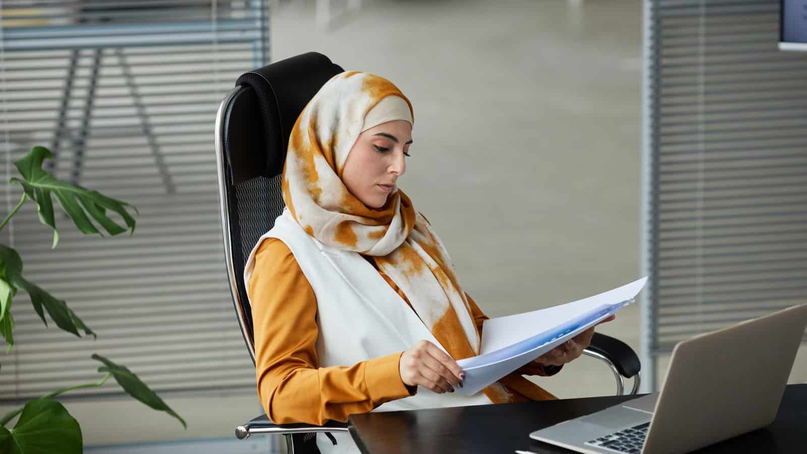 A woman in a hijab carefully reviewing documents at a desk with a laptop