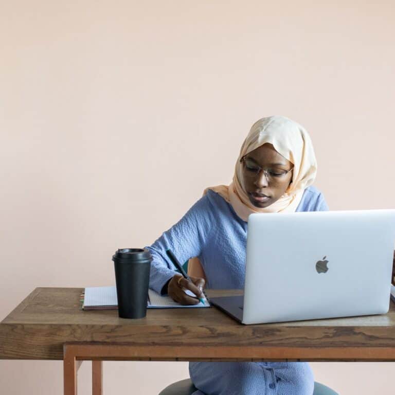 A woman in a peach hijab and long sleeves writing notes beside a laptop at a wooden desk