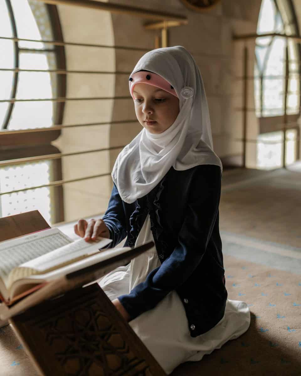 A young girl in a white hijab reading from a Quran on a wooden stand inside a mosque
