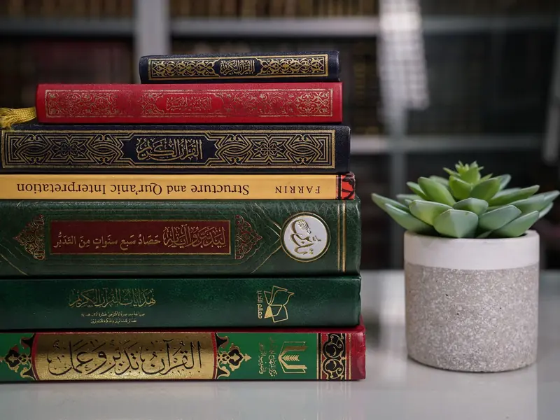 A stack of tafsir and Quran study books beside a small plant