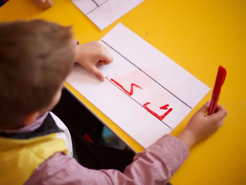 A young boy writing the Arabic letter kaf in red marker at a school desk