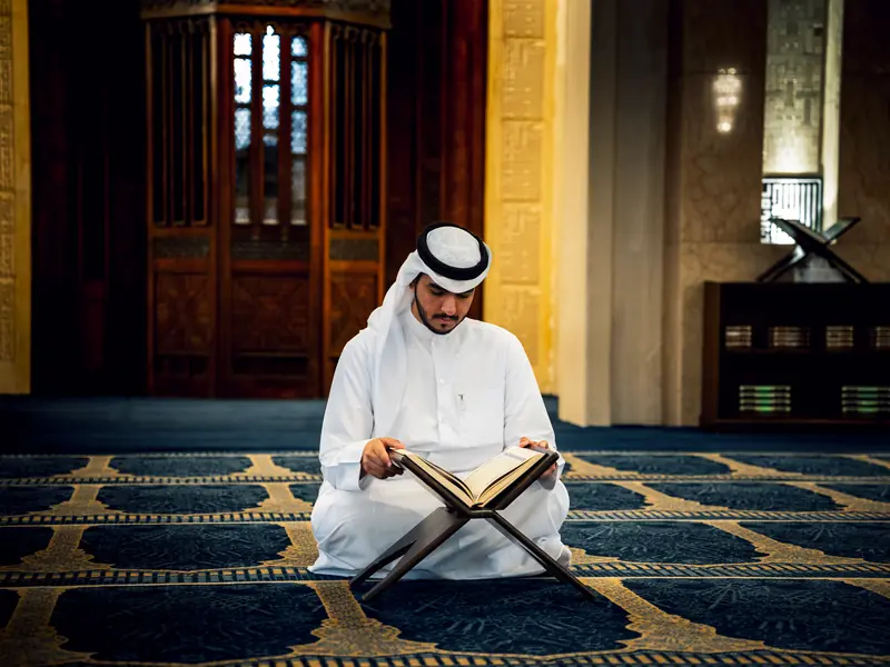 A man in a white thobe reading the Quran on a wooden stand in a mosque