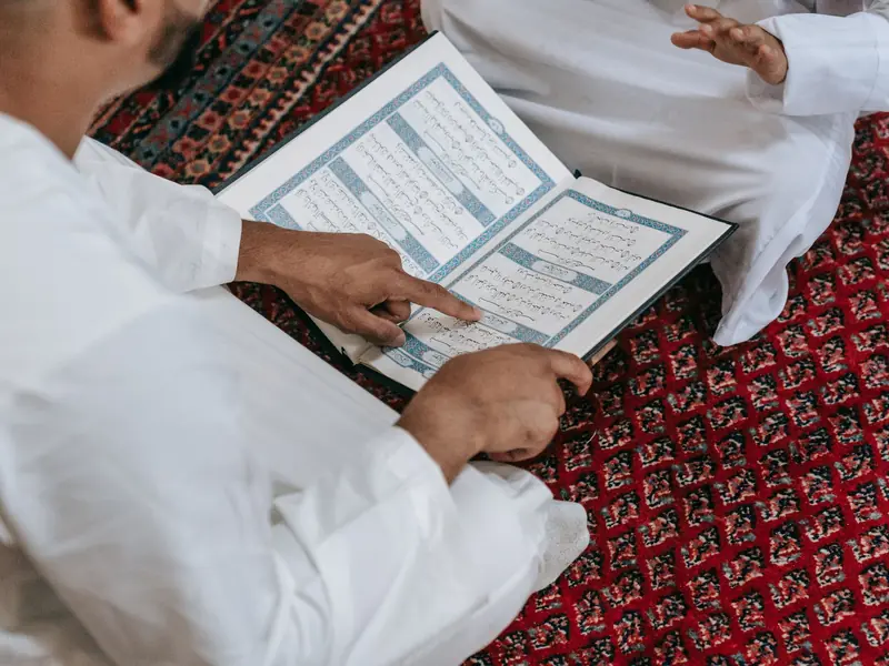 A teacher's finger guiding a student along a line of the Quran