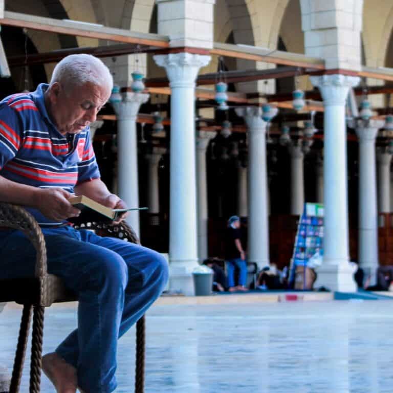 Elderly man reading a small Mushaf in the courtyard of a Cairo mosque, keeping up his Quran revision