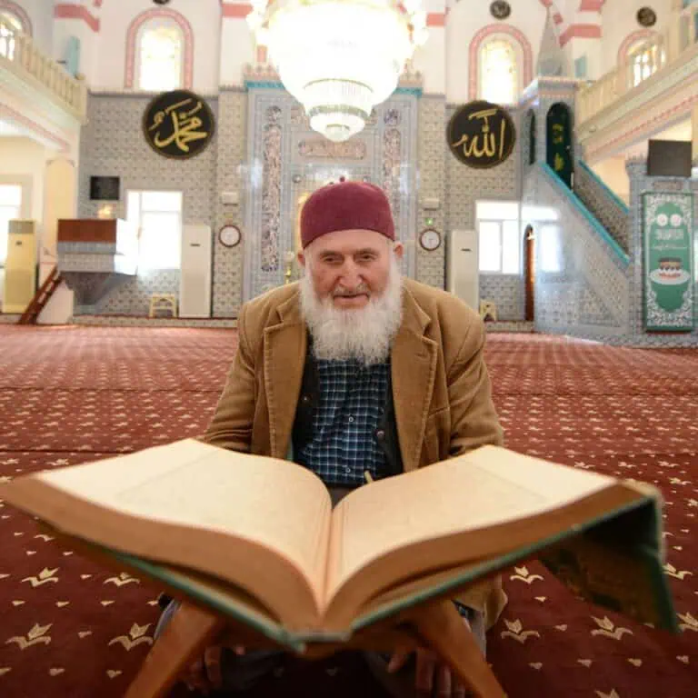 An elderly man with a white beard and a red kufi reading from a large Quran on a wooden stand in a mosque