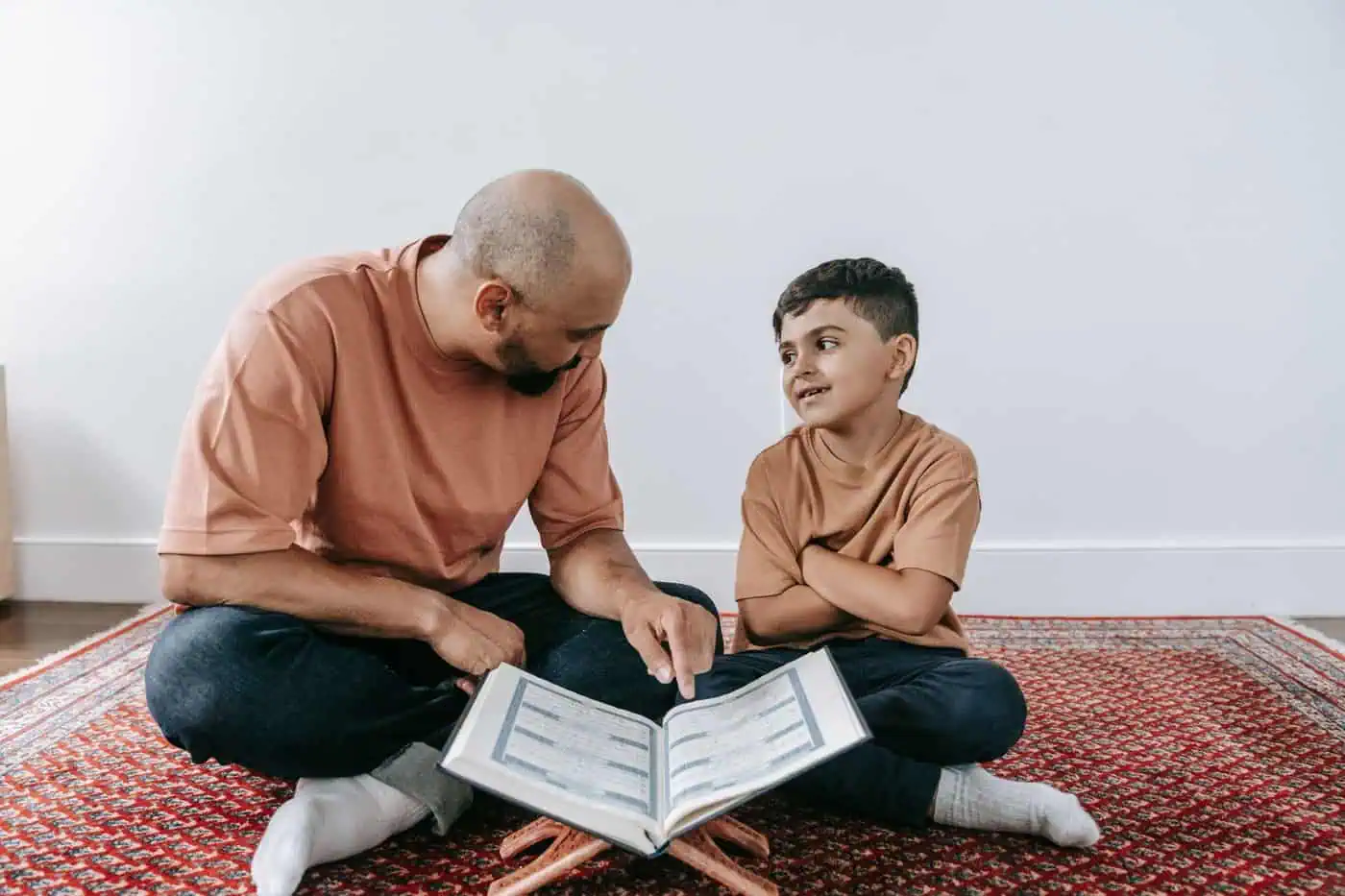 A father pointing to a line in a Quran on a wooden stand while his young son sits cross-legged beside him on a red rug